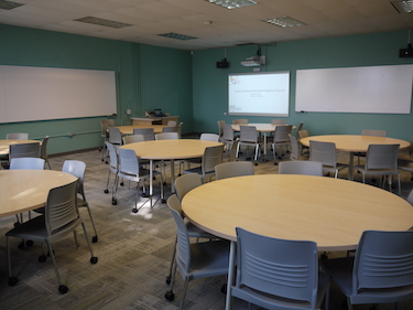 Classroom with round tables and movable chairs with whiteboards and a projection screen at the front of the room. 