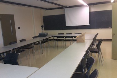 View from the side of the room: Narrow moveable tables with standard  moveable chairs set up in a rectangle with a blackboard on one wall.