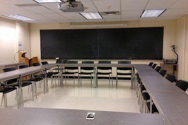 View from the back of the room: Narrow moveable tables with standard  moveable chairs set up in a rectangle with a whiteboard on one wall.