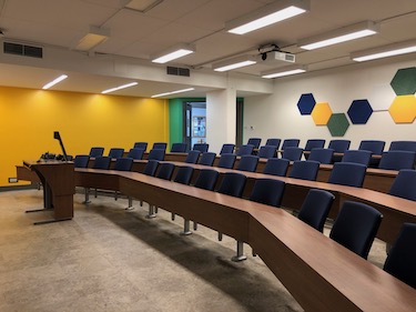 Classroom with 4 tiers of dark wood desks and blue chairs that swing out from desks. A bright yellow painted wall on the side of the classroom by the entry door. 