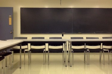 View from the back of the room: Narrow moveable tables with standard  moveable chairs set up in a rectangle with a blackboard on one wall.
