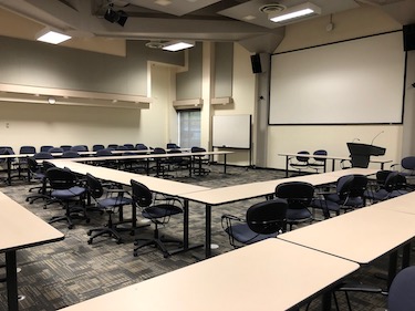 A large classroom with blue chairs and narrow tables arranged in a u-shape. A podium and screen are at the front of the room. 