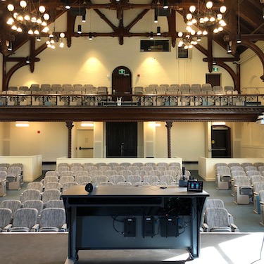 View from the front of the room: 2 level auditorium with rows of fixed chairs with tablet tables attached. The walls are an off white.