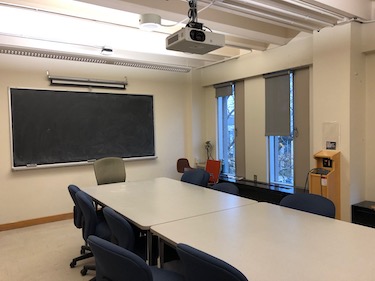 View from the side of the room: 2 large tables in the center of the room with moveable chairs surrounding them with a projector screen at the end of the room.