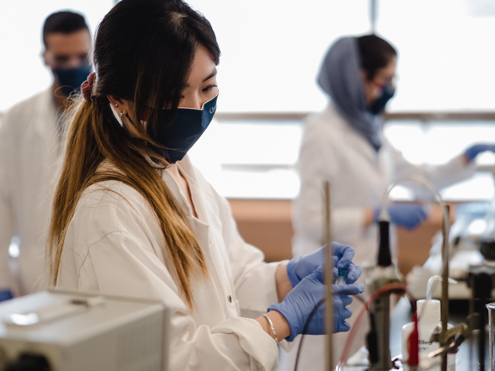 Masked woman in lab coat holding a test tube.