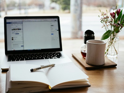 laptop with documents open and a notebook beside it with a pen resting on the open pages. There is a mug and honey beside the laptop