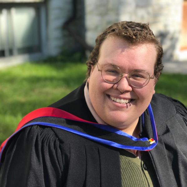 Picture of Wilde, fat person with short cropped brown hair wearing glasses.  They are wearing a Master of Philosophy hood and graduate gown, and there is a green sweater vest underneath the robe. There is green grass and a grey stone building behind them. 