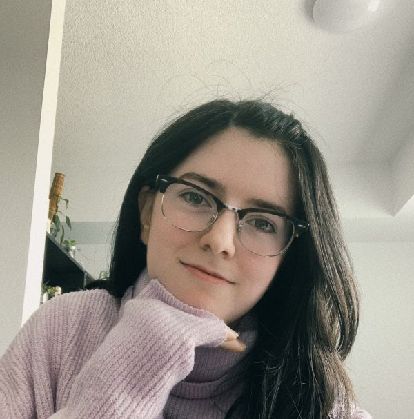 Photo of a young woman with brown hair and glasses. The photo displays her from the shoulders up smiling as her hand rests under her chin.