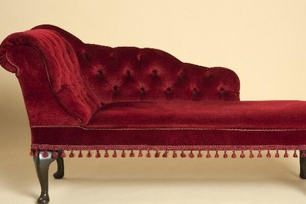 Photograph of a vintage-style chaise lounge upholstered in deep red velvet with tufted backrest and curved armrest on one side.