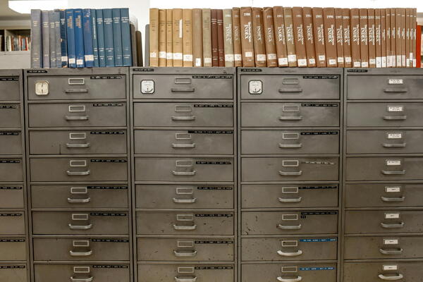 Photograph of multiple vintage metal filing cabinets with numerous drawers, topped with organized rows of hardcover books in blue, tan, and brown colors.