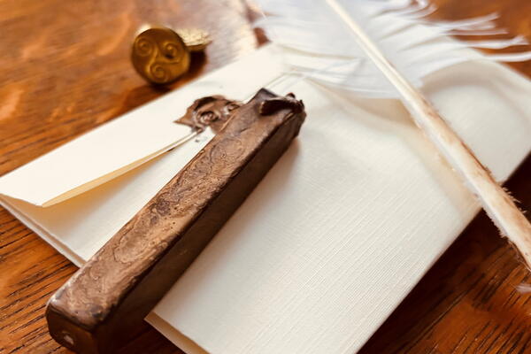 Photograph of vintage writing tools arranged on a wooden surface, including a white feather quill, sealed envelopes with wax stamps, and a smoking incense stick in a carved holder. The composition highlights historical communication methods with warm lighting emphasizing textures of paper, wax, and wood.