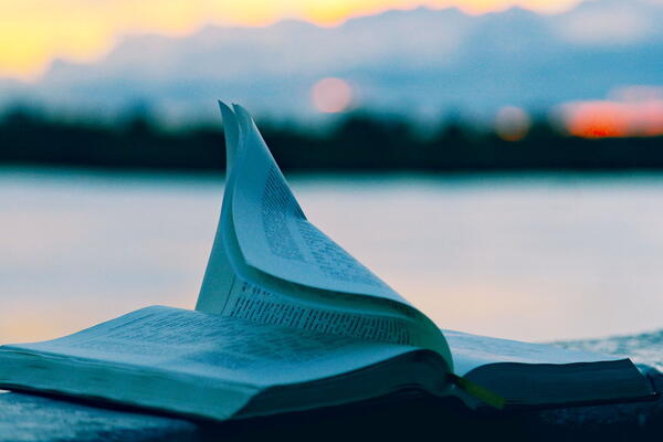Photograph of an open book with pages gently flipping, set against a blurred background of a body of water and distant mountains during sunset.