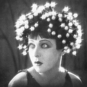 Black and white photograph of a person wearing a headdress made of numerous glowing orbs arranged in a circular pattern around their head. The image highlights the contrast between the bright lights and the dark background, creating a surreal and artistic effect.