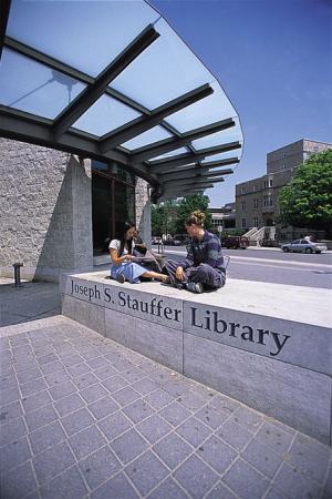 Students sitting on a stone sign outside; "Joseph S. Stauffer Library"