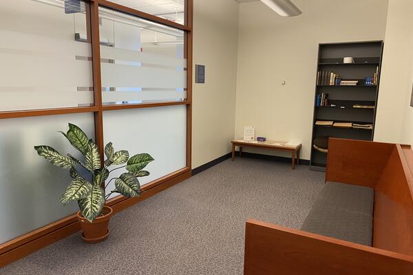 Stauffer Library room grey floors, book shelf to the left, seating area faces frosted glass wall and wide leafed potted plant on the floor. Go to campus map.