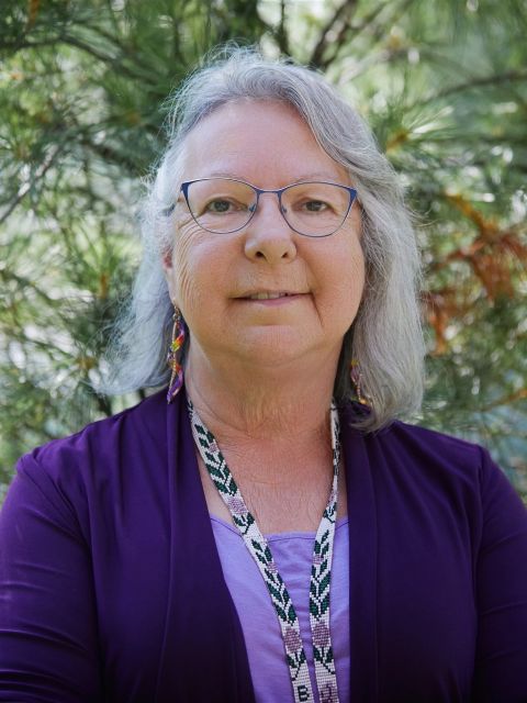 a photo of an older woman, an Indigenous elder in a purple top. she is wearing glasses and smiling, standing in front of a tree