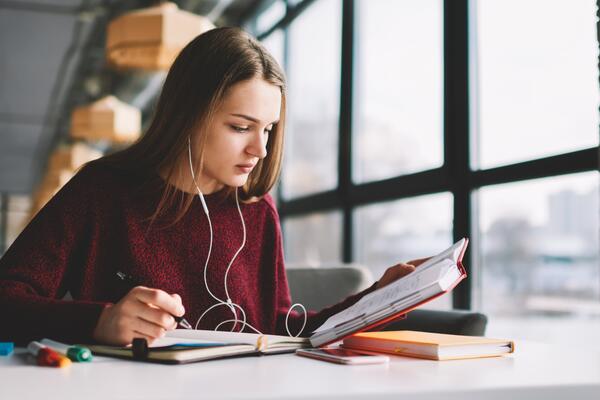 Pensive girl learning language online via earphones using application while writing new words into book