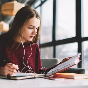 Pensive girl learning language online via earphones using application while writing new words into book