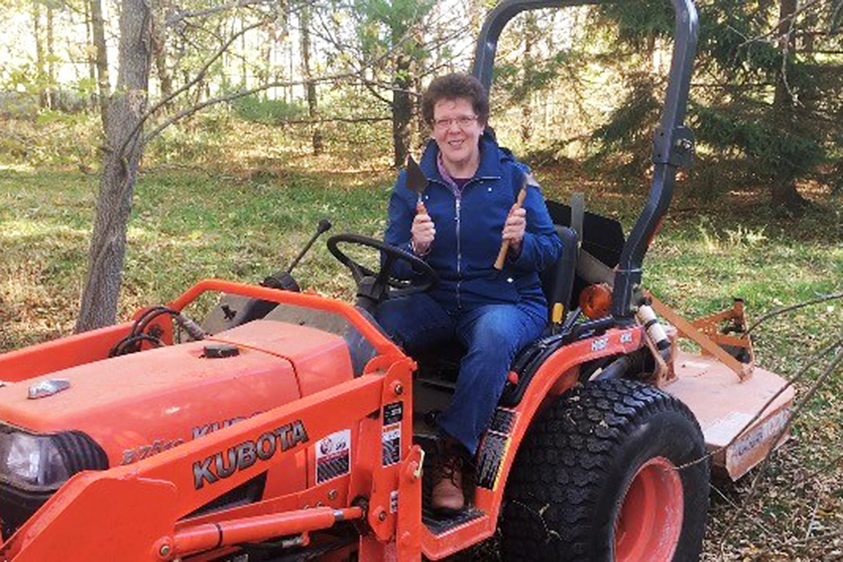 Classics professor Barbara Reeves holds excavating tools as she sits on an orange Kubota tractor.