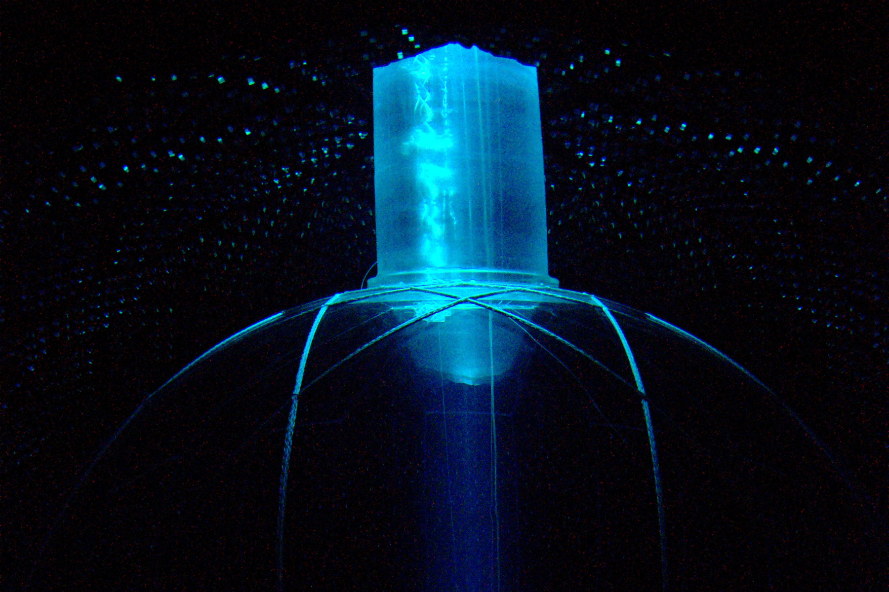 The detector consists of an active volume of 780 tonnes of liquid scintillator housed within a 12-metre diameter acrylic vessel that is held in place by ropes and viewed by an array of about 10,000 photomultiplier light detectors. In this image, taken by a camera embedded in the photomultiplier array, the detector is illuminated only by light from the clean room at the top of the vessel neck, producing a beam effect.