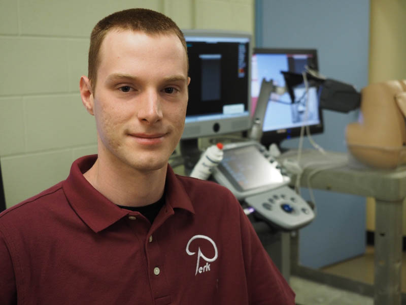 "Queen’s PhD student Matthew Holden sits in front of a microscope at the Laboratory for Percutaneous Surgery"