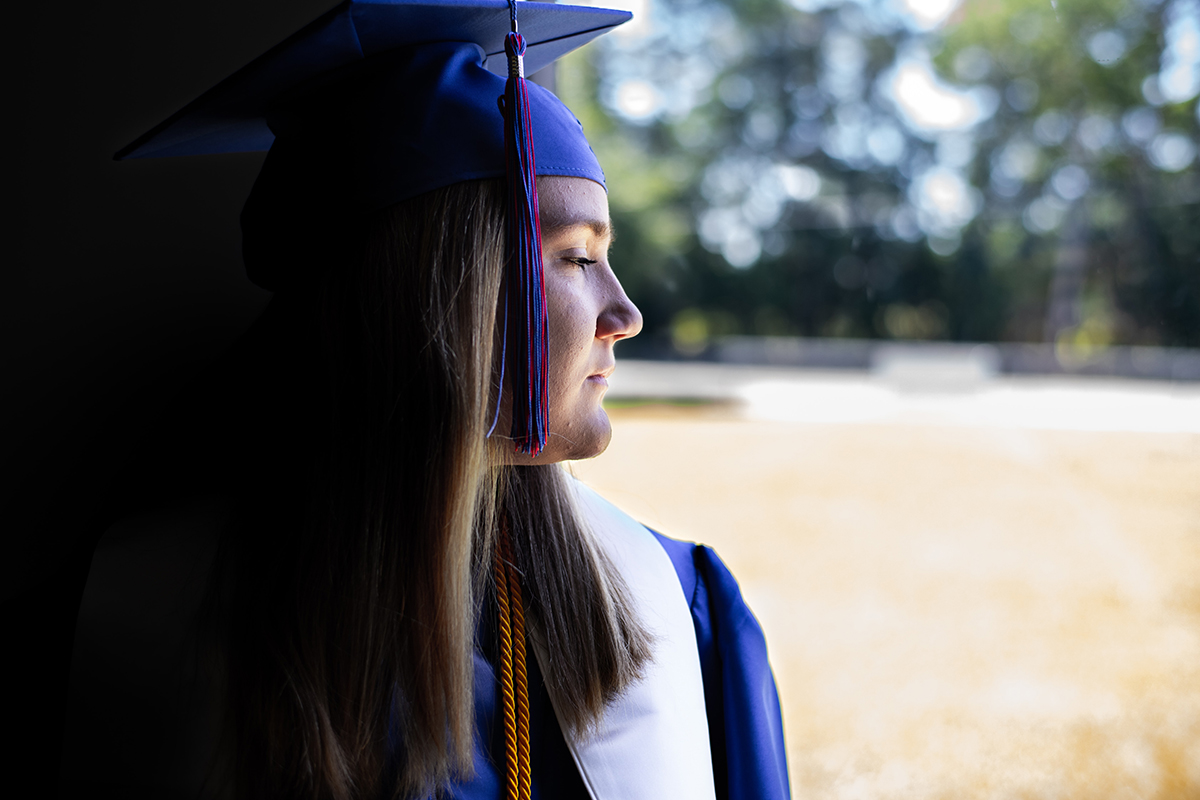 A lone high school graduate looks out onto an empty yard.
