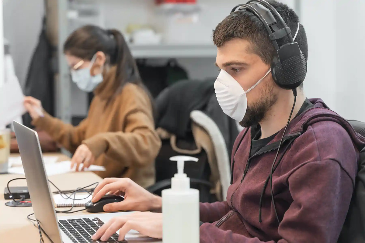 A young man with a mask works at a laptop.