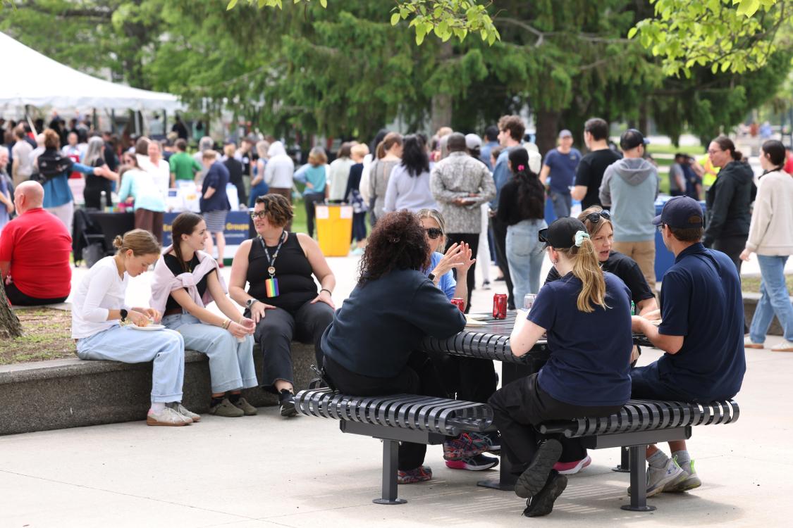 People sitting at a picnic table eating food and chatting, with large event tents and a lot of people in the background.