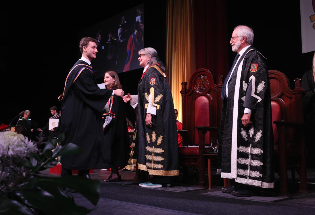 Young man walking across stage at convocation shaking hand with Shelagh Rogers.