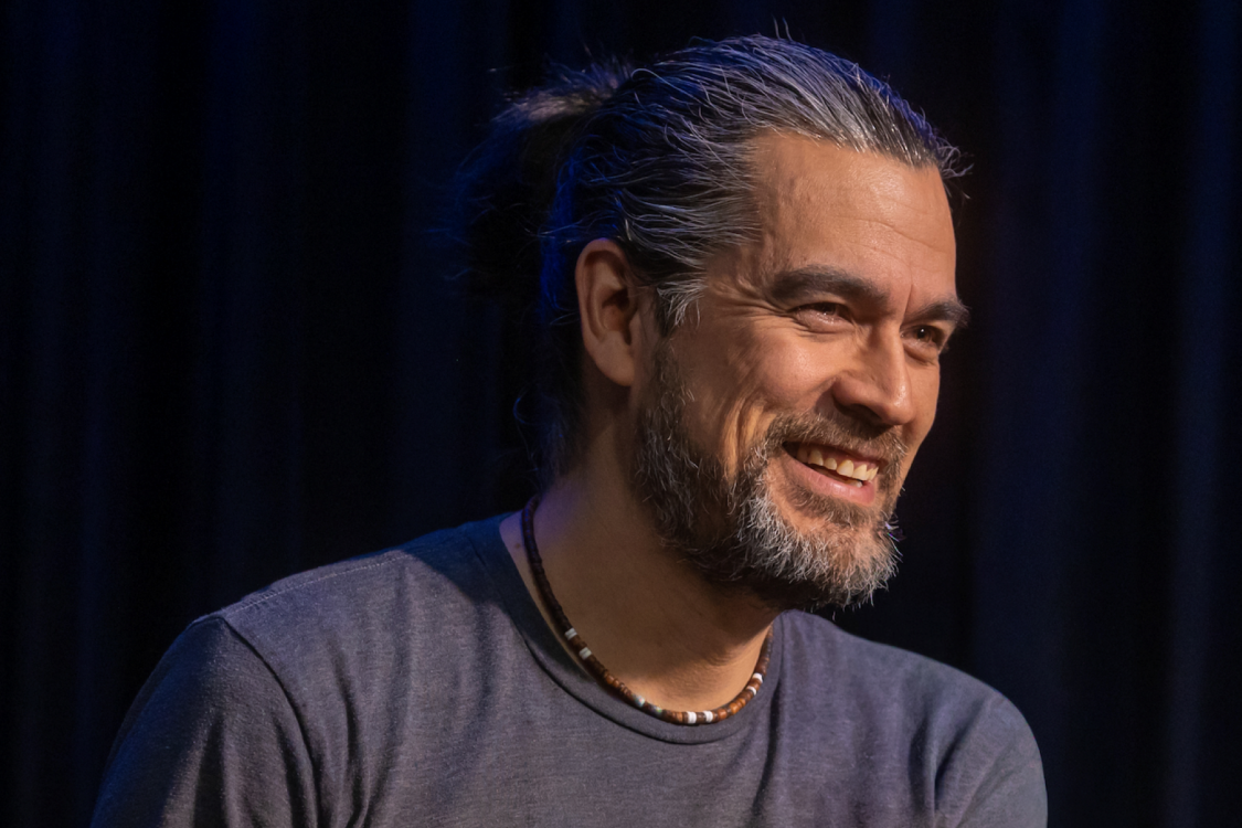 Man with long hair wearing dark grey t-shirt and beaded necklace smiling, in front of black background.
