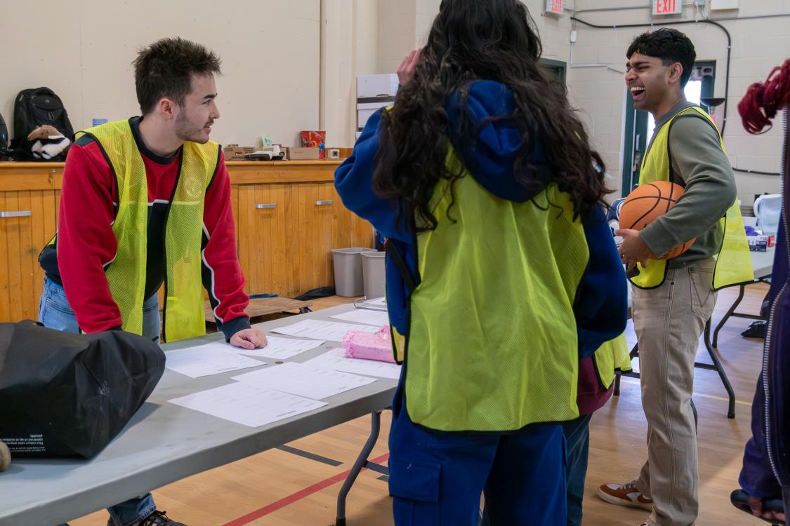 Three volunteers in yellow vests talking around a table in a gymnasium.