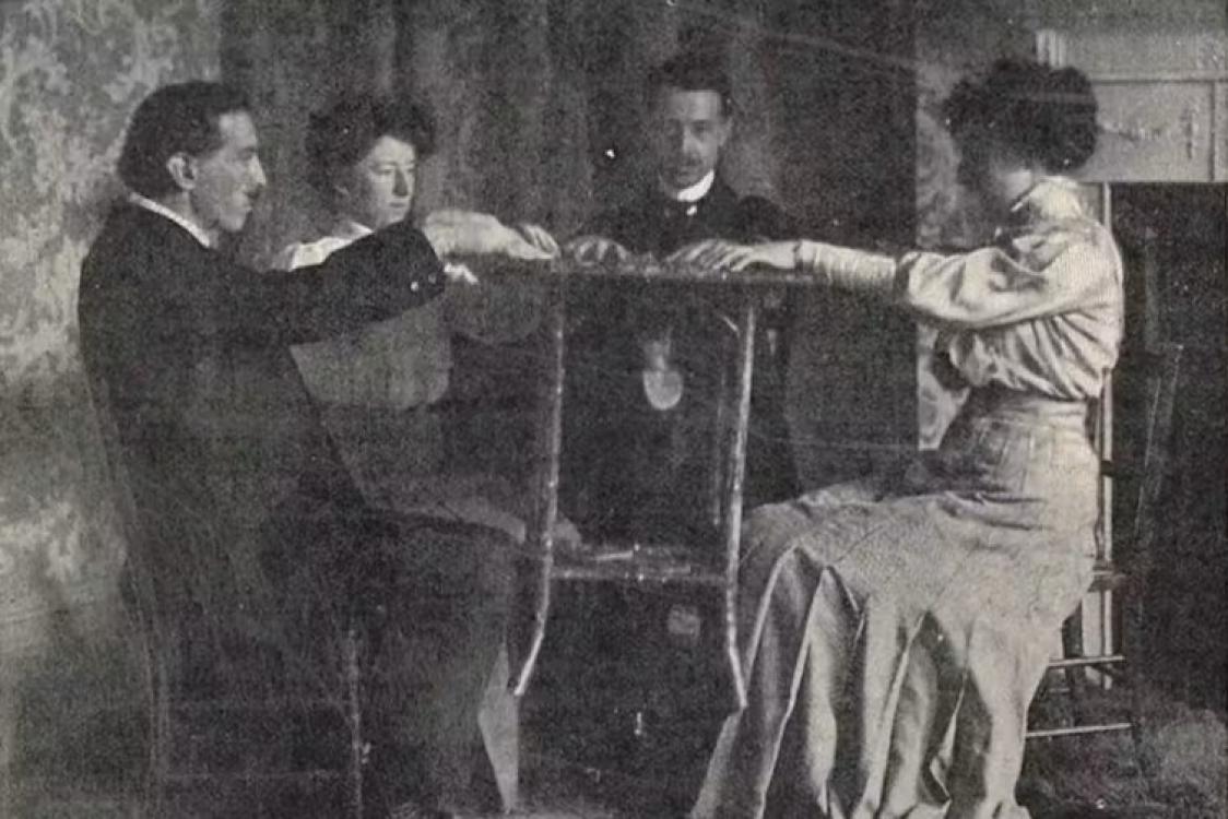 Three men and one woman siting in chairs facing each other, performing a séance.