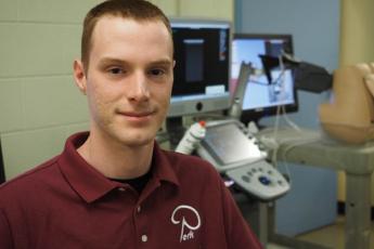 "Queen’s PhD student Matthew Holden sits in front of a microscope at the Laboratory for Percutaneous Surgery"