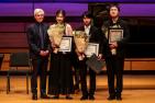 Older man in black suit standing with three young people (one woman and two men) who are holding awards and bouquets.