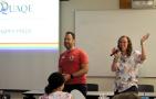 A man and a woman standing in the front of a classroom, beside a projector screen. The woman is speaking into a microphone.