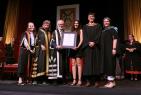 Six people standing on stage at a convocation standing beside one another, as one receives an award.