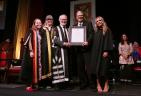 Five people standing on stage at a convocation standing beside one another, as one receives an award.