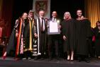 Six people standing on stage at a convocation standing beside one another, as one receives an award.