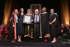 Six people standing on stage at a convocation standing beside one another, as one receives an award.