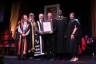 Six people standing on stage at a convocation standing beside one another, as one receives an award.
