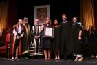 Seven people standing on stage at a convocation standing beside one another, as one receives an award.
