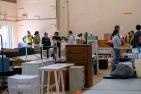 Group of people standing in a warehouse with donated furniture. Some are volunteers wearing yellow vests.