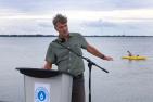 Man speaking at podium on pier, pointing to a kayak in the water.