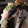 Woman in white dress, holding a handkerchief, dancing in front of a background of balloons.
