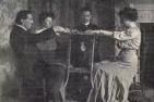 Three men and one woman siting in chairs facing each other, performing a séance.