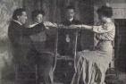 Three men and one woman siting in chairs facing each other, performing a séance.
