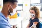 A young girl wearing a mask is vaccinated by a male doctor wearing a surgical mask.