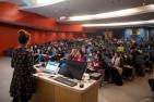 A professor delivers a class in a lecture hall with red walls in background.