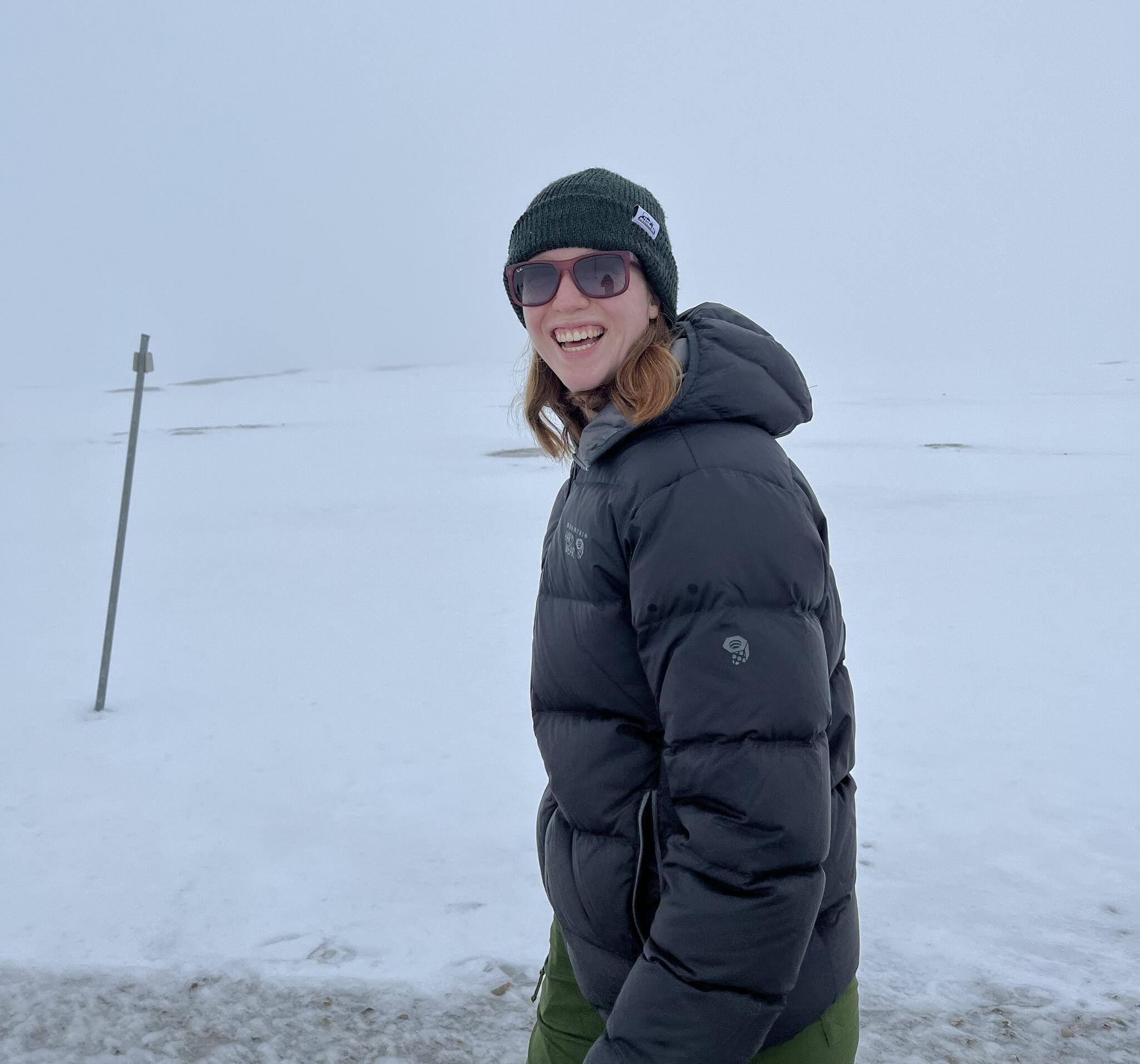 Gillian standing in a snowy environment, smiling, wearing sunglasses, a black winter coat, a green hat, and green pants. 