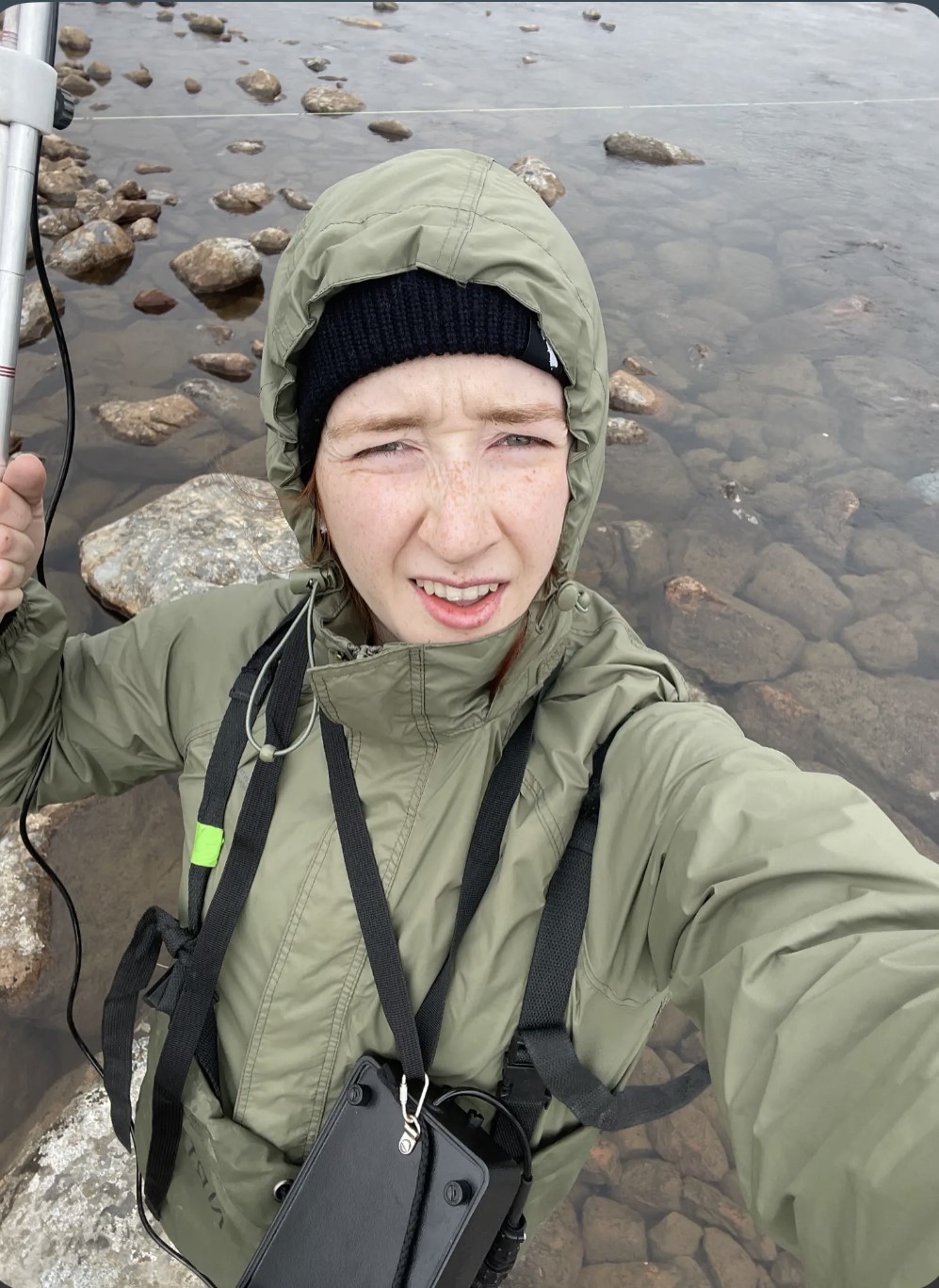 Selfie of Grace Ayers, standing in a shallow pool of water with visible large stones under approximately 10cm of water. Grace is wearing an olive green rain jacket with the hood pulled up and has a black toque underneath. Grace is holding a piece of equipment (a silver colored rod) just out of view on the left of the image. Grace is carrying a small black bag across their front.
